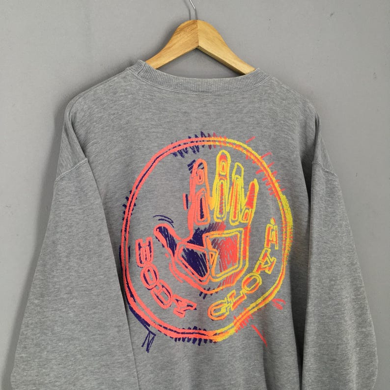 May include: Gray crewneck sweatshirt with a colorful graphic on the back. The graphic features a hand inside a circle with the words "BODY GLOVE" in a neon orange, yellow, and purple design. The sweatshirt is hanging on a wooden hanger.