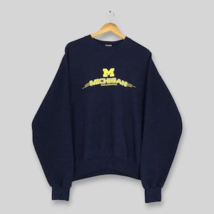 May include: A navy blue crewneck sweatshirt featuring the yellow Michigan Wolverines logo and text printed on the chest. The garment has long sleeves with ribbed cuffs and a ribbed hem, and it is displayed on a wooden hanger against a plain background.