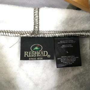 Peut inclure: Gros plan sur une &eacute;tiquette de la marque RedHead sur un sweat &agrave; capuche. L'&eacute;tiquette noire porte du texte blanc, le logo RedHead et l'inscription "SINCE 1856". Des instructions de lavage et la composition du tissu sont &eacute;galement visibles.