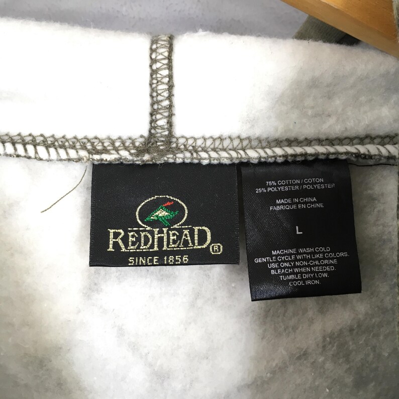 Peut inclure: Gros plan sur une &eacute;tiquette de la marque RedHead sur un sweat &agrave; capuche. L'&eacute;tiquette noire porte du texte blanc, le logo RedHead et l'inscription "SINCE 1856". Des instructions de lavage et la composition du tissu sont &eacute;galement visibles.