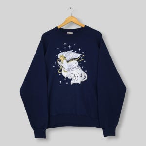 May include: A navy blue long-sleeve sweatshirt featuring a central graphic of an angel with flowing white robes and wings, surrounded by small white stars. The angel holds a golden ribbon, and the design is printed on the front of the garment.
