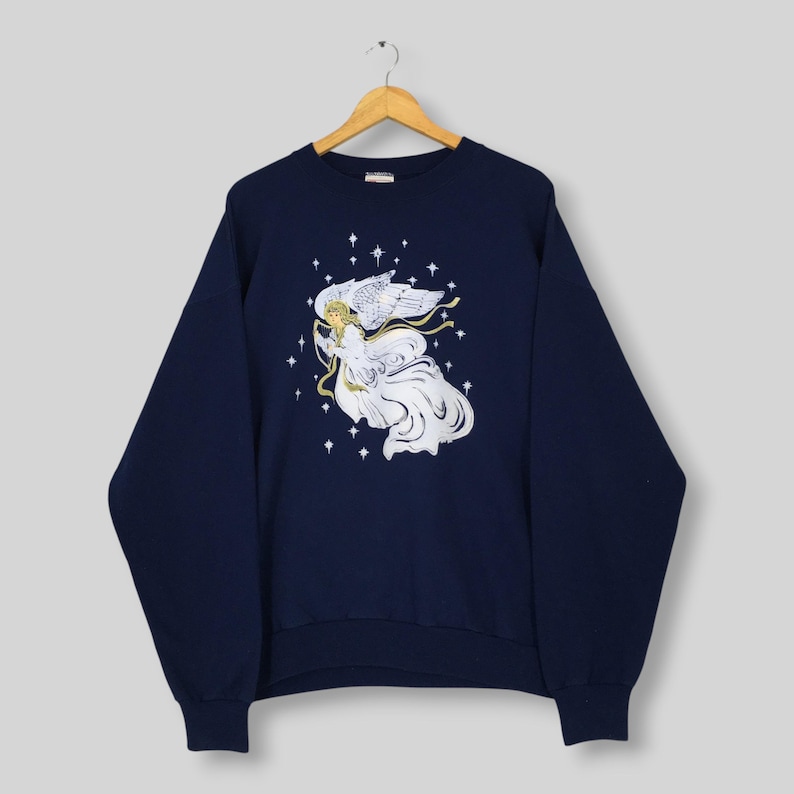 May include: A navy blue long-sleeve sweatshirt featuring a central graphic of an angel with flowing white robes and wings, surrounded by small white stars. The angel holds a golden ribbon, and the design is printed on the front of the garment.