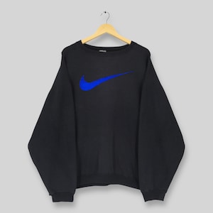Op de afbeelding: Zwart sweatshirt met ronde hals en een groot blauw Nike swoosh-logo op de voorkant. Het sweatshirt hangt aan een houten hanger tegen een lichtgrijze achtergrond. Het logo is helderblauw.