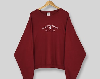May include: A maroon crewneck sweatshirt featuring the white embroidered text Indiana University and Hoosiers with a central IU logo. The long-sleeve pullover has a ribbed neckline, cuffs, and hem, and is displayed on a wooden clothes hanger.