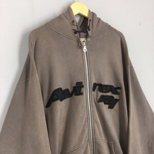 Puede incluir: Una sudadera con capucha marr&oacute;n con cremallera y el texto "Avi Rex Ny" en letras negras. La sudadera con capucha tiene una capucha con cord&oacute;n y una cremallera de largo completo. La prenda est&aacute; colgada en una percha de madera.