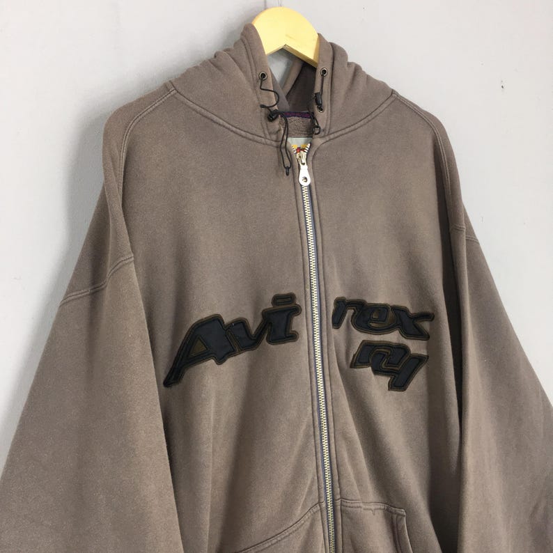 Puede incluir: Una sudadera con capucha marr&oacute;n con cremallera y el texto "Avi Rex Ny" en letras negras. La sudadera con capucha tiene una capucha con cord&oacute;n y una cremallera de largo completo. La prenda est&aacute; colgada en una percha de madera.