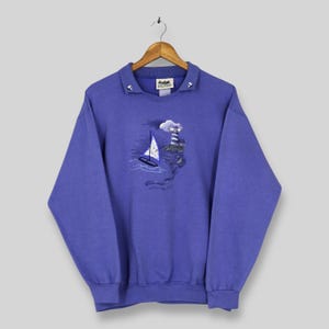 Könnte beinhalten: Ein periwinkelfarbenes Sweatshirt mit Kragen und nautischem Design. Die gestickte Grafik zeigt ein Segelboot, einen Leuchtturm und Meereswellen. Das Sweatshirt hängt an einem Holzkleiderbügel.