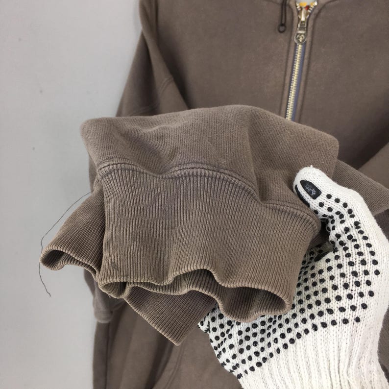 Puede incluir: Primer plano de una sudadera con capucha marr&oacute;n con cremallera. El pu&ntilde;o acanalado de la manga es visible, con una ligera decoloraci&oacute;n. La cremallera es plateada con un tirador dorado. La sudadera con capucha est&aacute; sujeta por una mano enguantada.