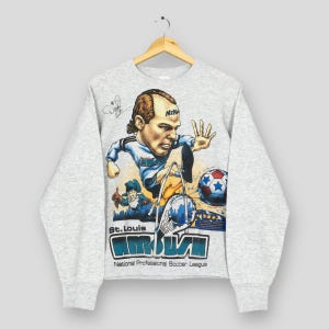 Puede incluir: Sudadera gris claro con una ilustración de dibujos animados de un jugador de fútbol. El diseño incluye el texto "St. Louis AMBUSH" y "National Professional Soccer League". El logotipo de Nike es visible.