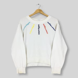 May include: White sweatshirt with the word "courr&egrave;ges" embroidered in blue, yellow, and red. The text is arranged in a fan shape across the chest. The sweatshirt has long sleeves and a crew neck.