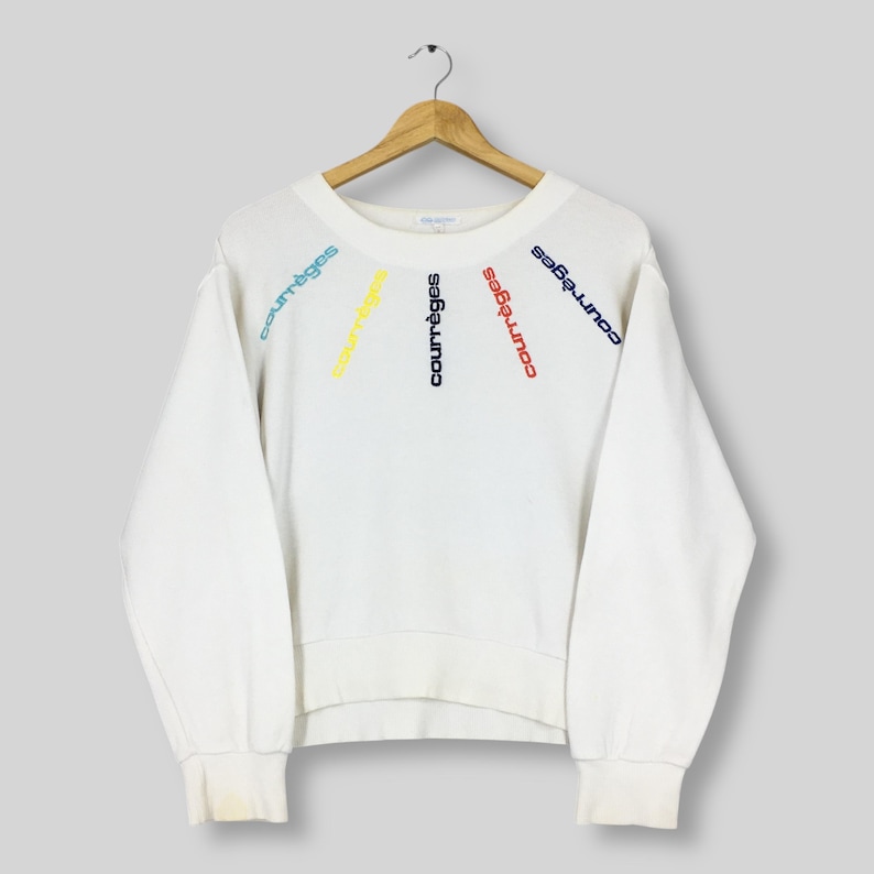 May include: White sweatshirt with the word "courr&egrave;ges" embroidered in blue, yellow, and red. The text is arranged in a fan shape across the chest. The sweatshirt has long sleeves and a crew neck.