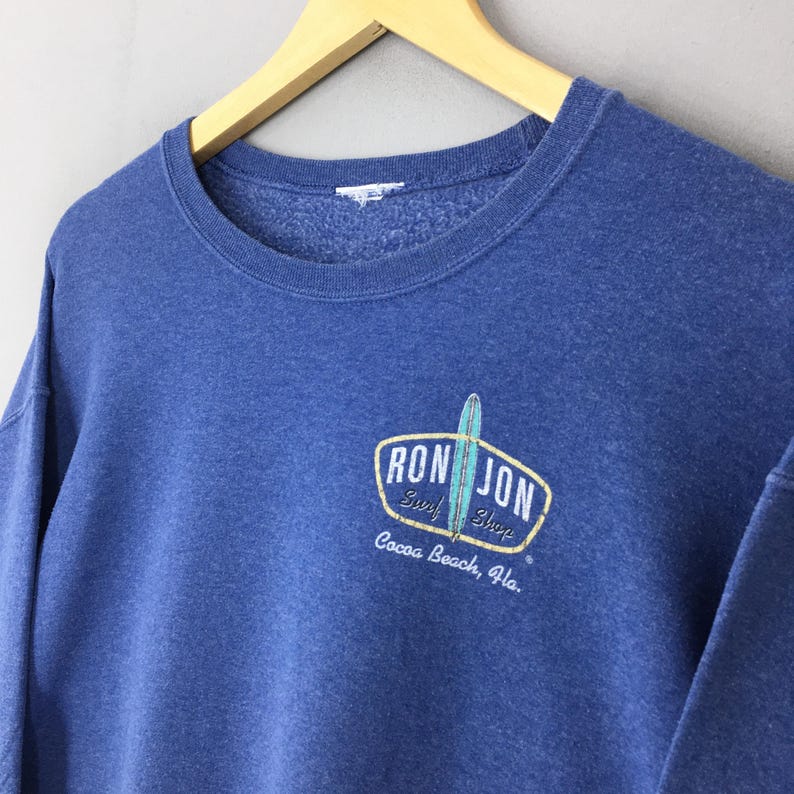 May include: A blue crewneck sweatshirt featuring a graphic on the chest with the text RON JON Surf Shop Cocoa Beach, Fla. and an illustration of a surfboard. The sweatshirt has a ribbed collar and a soft, textured interior lining.
