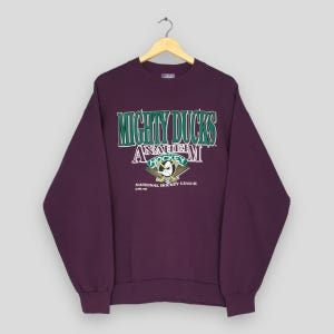 Op de afbeelding: Een donkerpaarse sweatshirt met de tekst "MIGHTY DUCKS" in groene en witte letters. Daaronder staat "ANAHEIM HOCKEY" met een eendenlogo en gekruiste hockeysticks. De tekst "NATIONAL HOCKEY LEAGUE" staat eronder.