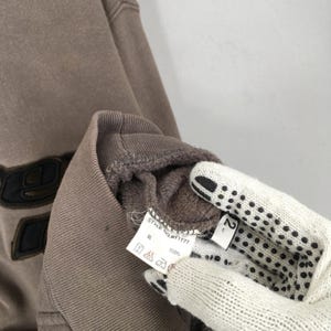 Puede incluir: Primer plano de una sudadera marr&oacute;n con un dise&ntilde;o negro en la parte delantera. La prenda tiene cuello y pu&ntilde;os acanalados. Se ve una etiqueta con el texto "STYLE no. 01777" y s&iacute;mbolos de cuidado.