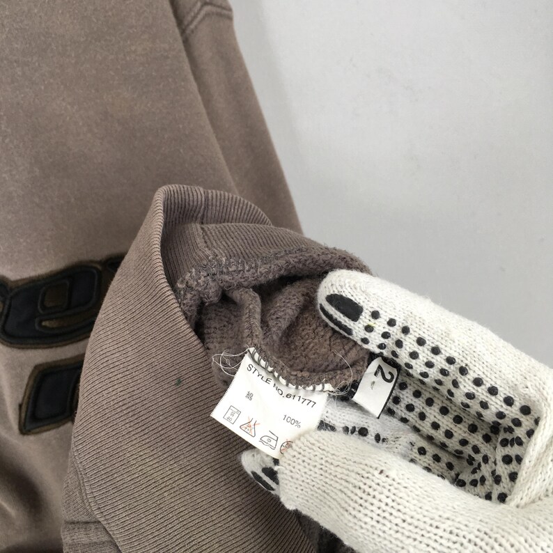 Puede incluir: Primer plano de una sudadera marr&oacute;n con un dise&ntilde;o negro en la parte delantera. La prenda tiene cuello y pu&ntilde;os acanalados. Se ve una etiqueta con el texto "STYLE no. 01777" y s&iacute;mbolos de cuidado.