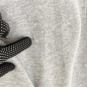 May include: Close-up of a light gray fabric with a small tear. The fabric has a textured appearance, and the tear reveals the inner threads. The image is well-lit, highlighting the details of the material.