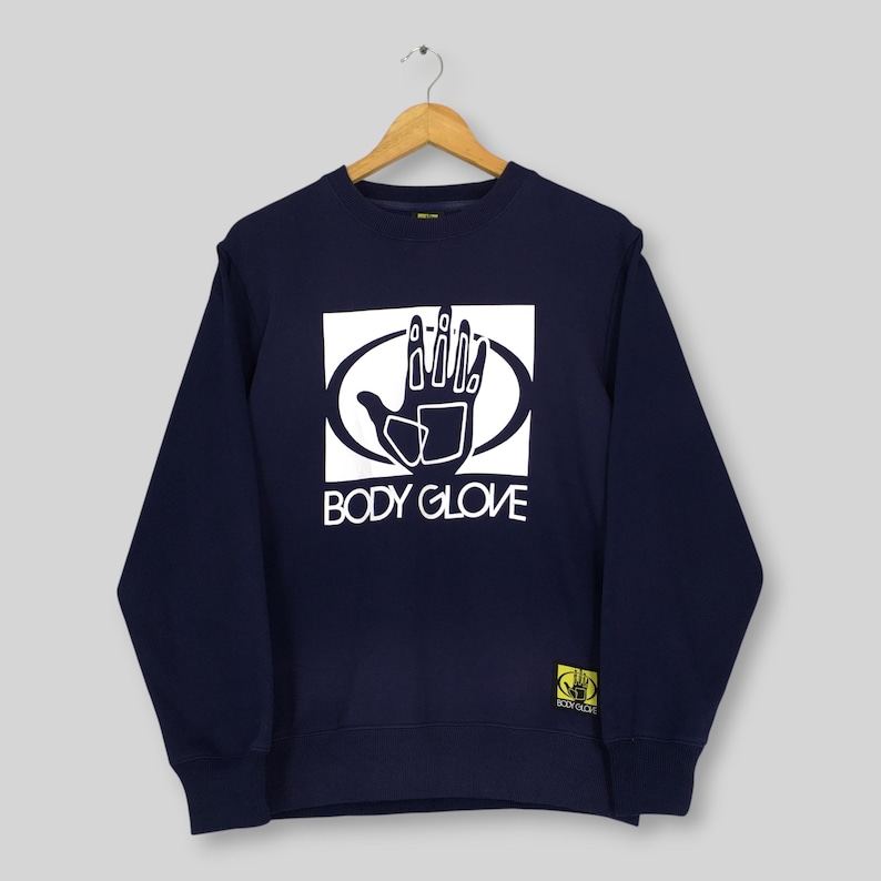May include: A navy blue long-sleeve sweatshirt featuring a white graphic of a hand inside an oval shape with the text BODY GLOVE printed below. A small yellow rectangular patch with a black hand logo is located at the bottom left corner.
