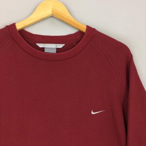 Może przedstawiać: Bordowa bluza z okrągłym dekoltem i małym białym logo Nike na lewej piersi. Bluza wisi na drewnianym wieszaku. Materiał wydaje się być miękką, wygodną tkaniną.