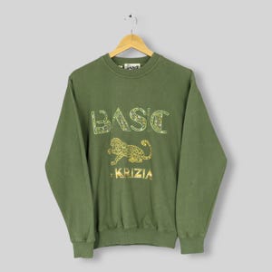 May include: Olive green sweatshirt with the word "BASIC" in a decorative font and a gold leopard graphic. The brand name "KRIZIA" is printed below the leopard. The sweatshirt is hanging on a wooden hanger.