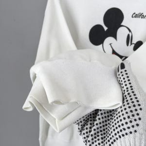 May include: White sweatshirt featuring a black and red Mickey Mouse graphic. The sweatshirt has a crew neck and long sleeves with ribbed cuffs. The sleeve is being held by a white glove with black polka dots. The word "Califor" is visible.