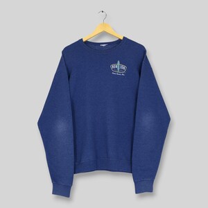 May include: A navy blue crewneck sweatshirt featuring long sleeves and a ribbed collar, cuffs, and hem. On the upper left chest, there is a small graphic logo that reads RON JON SURF SHOP with an illustration of a surfboard in the center.