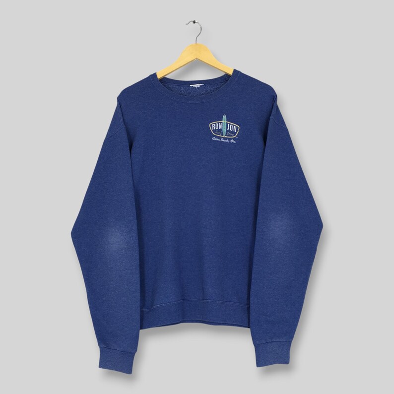May include: A navy blue crewneck sweatshirt featuring long sleeves and a ribbed collar, cuffs, and hem. On the upper left chest, there is a small graphic logo that reads RON JON SURF SHOP with an illustration of a surfboard in the center.