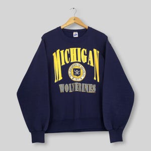 May include: A navy blue crewneck sweatshirt featuring the word MICHIGAN in large yellow letters arched over a circular University of Michigan seal. Below the seal, the word WOLVERINES is printed in grey block letters on the chest area.
