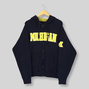 May include: A navy blue zip-up hoodie featuring the word MICHIGAN printed in bold yellow letters across the chest. The garment includes a drawstring hood with a yellow interior lining, a full-length front zip, and a yellow letter A on the sleeve.