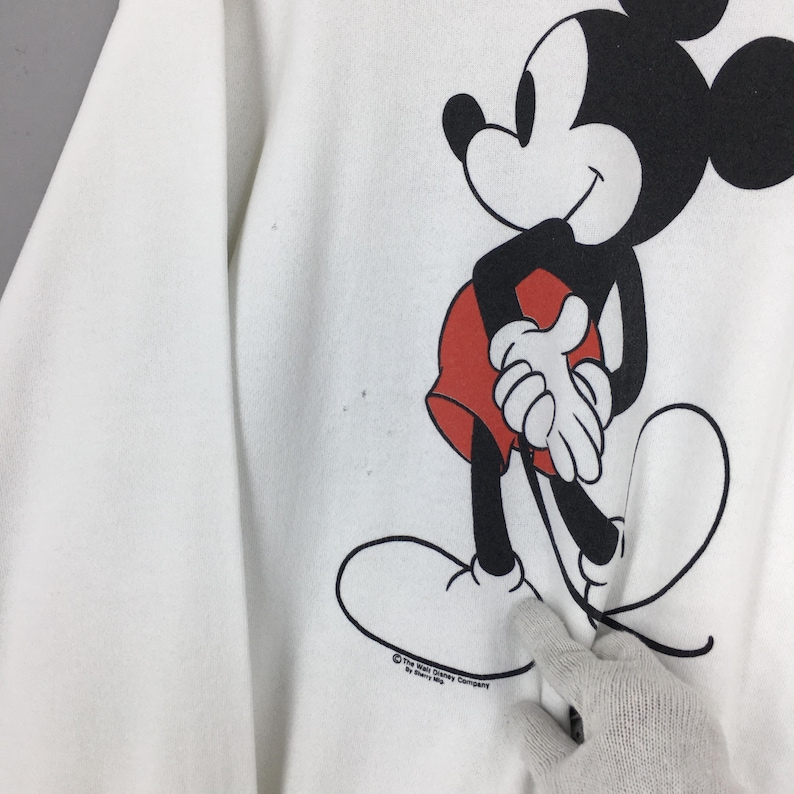May include: White sweatshirt featuring a large graphic of Mickey Mouse in black, red, and white. The cartoon character is smiling and standing with his hands clasped. The Walt Disney Company logo is printed at the bottom.