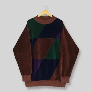 May include: An oversized brown sweatshirt with a geometric pattern in shades of brown, green, navy, and purple. The long sleeves and collar are a solid brown. The sweatshirt is hanging on a wooden hanger.
