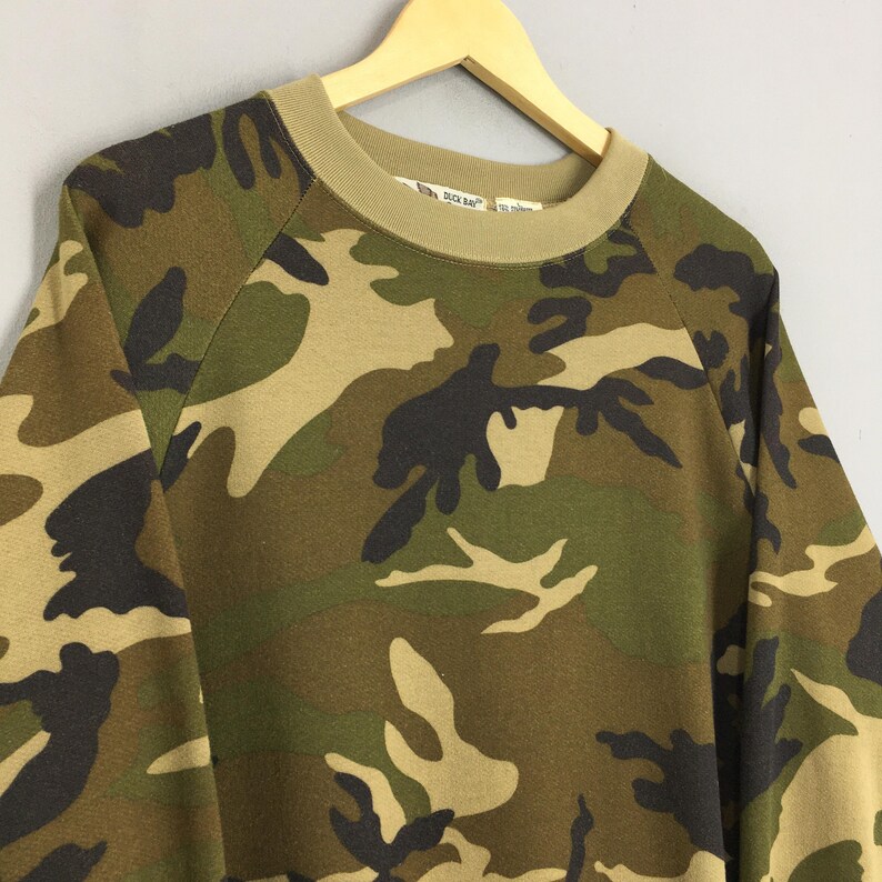 May include: Camouflage-patterned sweatshirt in shades of green, brown, and tan. The long-sleeved top features a crew neck and a relaxed fit. The sweatshirt is hanging on a wooden hanger.
