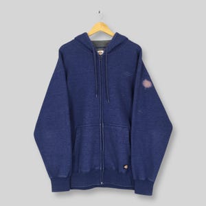 Könnte beinhalten: Ein marineblauer Kapuzenpullover mit Reißverschluss, Kordelzugkapuze und Vordertasche. Der Hoodie hat ein kleines Logo auf der Brust und ein größeres Logo unten rechts. Der Stoff hat ein verwaschenes Aussehen, und es gibt einen Fleck auf dem rechten Ärmel.