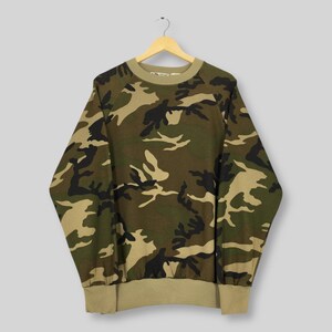 May include: A camouflage-patterned sweatshirt in shades of green, brown, and black hangs from a wooden hanger. The long-sleeved top features a crew neck and ribbed cuffs and hem in a solid tan color. The fabric displays a classic military-style print.
