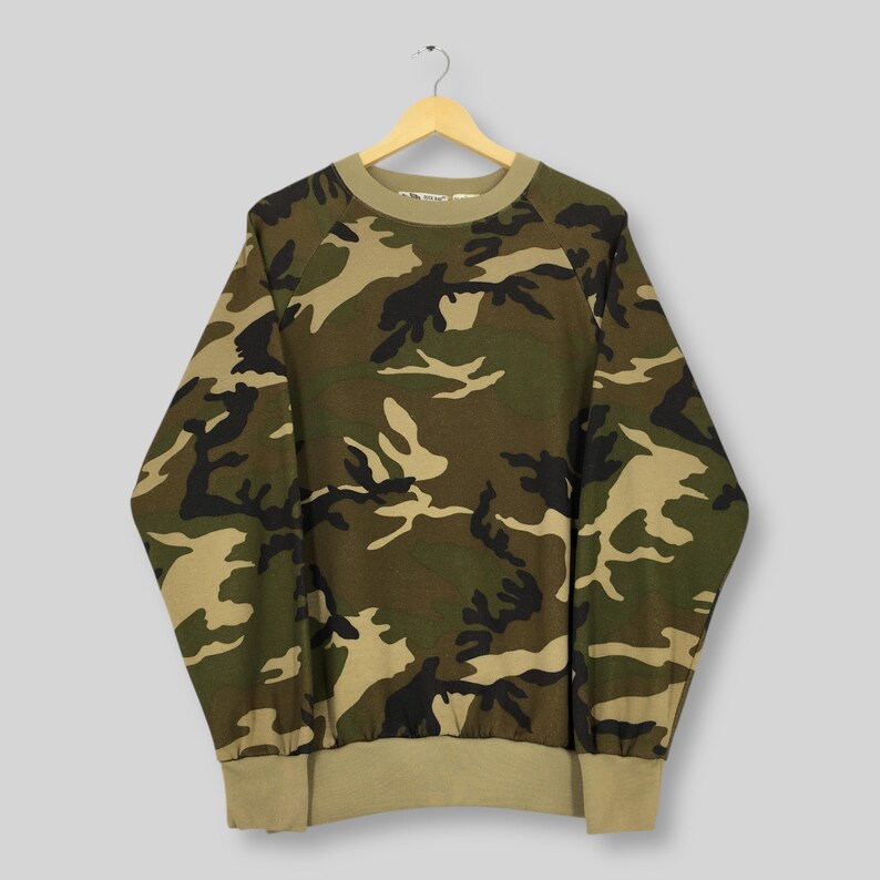 May include: A camouflage-patterned sweatshirt in shades of green, brown, and black hangs from a wooden hanger. The long-sleeved top features a crew neck and ribbed cuffs and hem in a solid tan color. The fabric displays a classic military-style print.