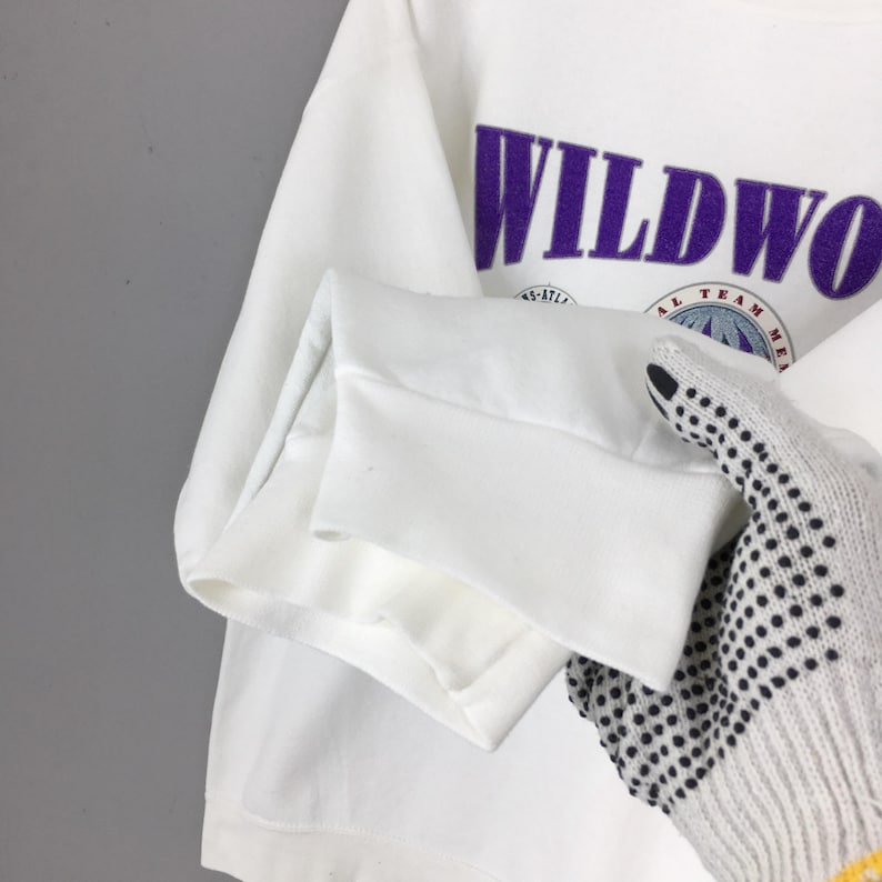 May include: White long-sleeved sweatshirt with the word "WILDWOOD" in purple block letters. Below the text is a circular graphic with the words "ATL TEAM ME". The sleeve cuff is visible.