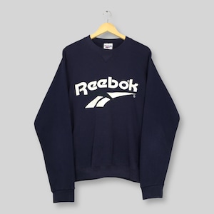 Op de afbeelding: Marineblauw Reebok sweatshirt met het witte logo van het merk op de borst. Het sweatshirt hangt aan een houten hanger tegen een lichte achtergrond. De top met lange mouwen heeft een ronde hals.