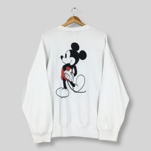 May include: White sweatshirt featuring a large graphic of Mickey Mouse on the back. The cartoon character is depicted in black, red, and white. The sweatshirt is hanging on a wooden hanger.