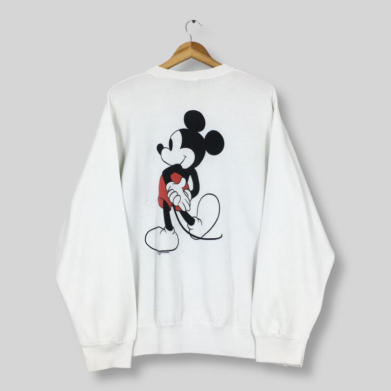 May include: White sweatshirt featuring a large graphic of Mickey Mouse on the back. The cartoon character is depicted in black, red, and white. The sweatshirt is hanging on a wooden hanger.