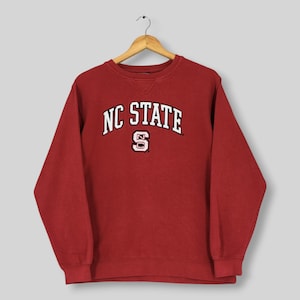 May include: A dark red crewneck sweatshirt featuring the text NC STATE in white arched lettering above a white and red block letter S logo. The garment has long sleeves, a ribbed collar with a V-shaped stitch detail, and ribbed cuffs.