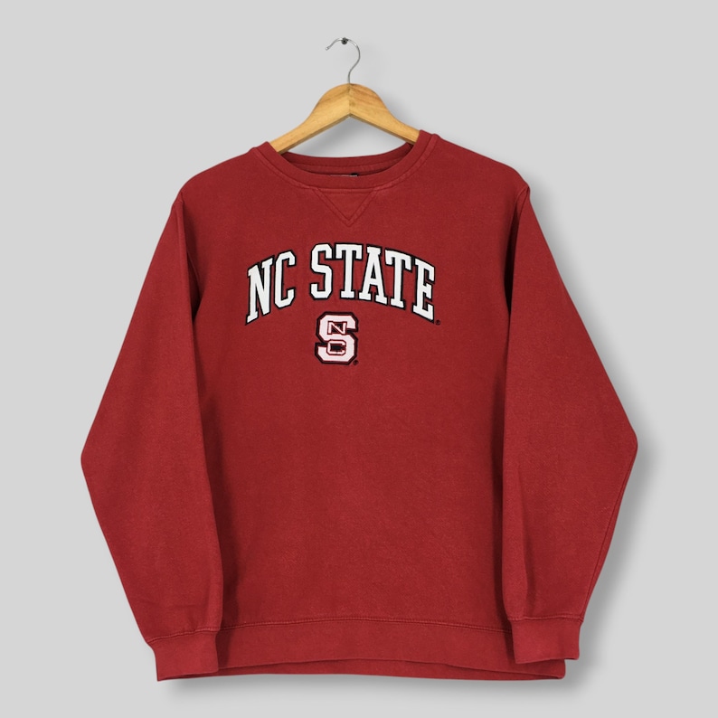 May include: A dark red crewneck sweatshirt featuring the text NC STATE in white arched lettering above a white and red block letter S logo. The garment has long sleeves, a ribbed collar with a V-shaped stitch detail, and ribbed cuffs.