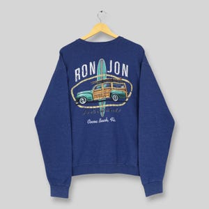 May include: A navy blue long-sleeve sweatshirt featuring a graphic on the back. The design includes a teal surfboard, a wood-paneled station wagon, and the text "RON JON" above, with "It's all about the surf!" and "Cocoa Beach, Fla." printed below.