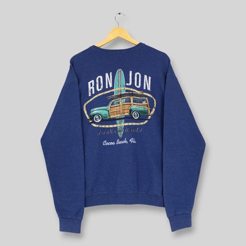 May include: A navy blue long-sleeve sweatshirt featuring a graphic on the back. The design includes a teal surfboard, a wood-paneled station wagon, and the text "RON JON" above, with "It's all about the surf!" and "Cocoa Beach, Fla." printed below.