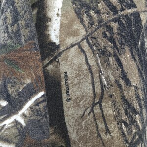 Op de afbeelding: Close-up van een stof met een Realtree camouflagepatroon. Het patroon heeft tinten bruin, groen en zwart, die lijken op een bosrijke omgeving. Het woord "Realtree" staat op de stof gedrukt.