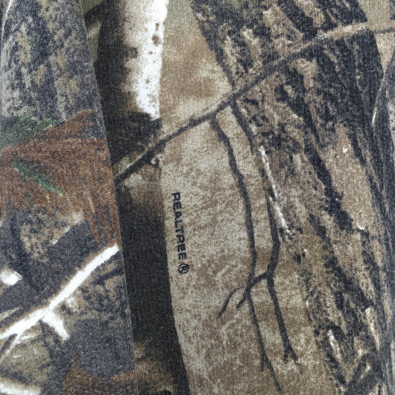Op de afbeelding: Close-up van een stof met een Realtree camouflagepatroon. Het patroon heeft tinten bruin, groen en zwart, die lijken op een bosrijke omgeving. Het woord "Realtree" staat op de stof gedrukt.