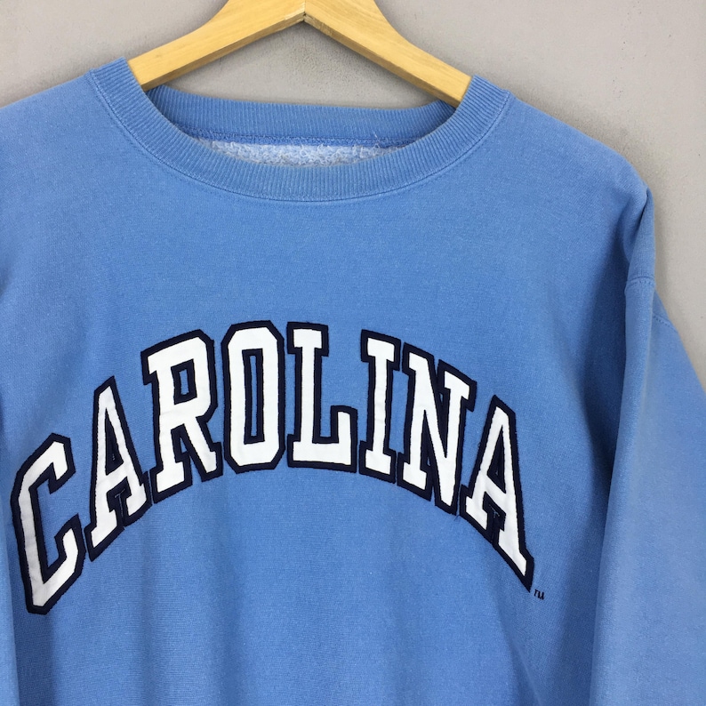 May include: A pale blue sweatshirt featuring the word "CAROLINA" in white, outlined in navy, across the chest. The garment is displayed on a wooden hanger.