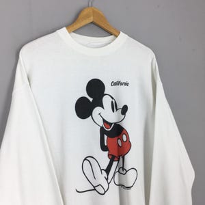 May include: White long-sleeve shirt featuring a large graphic of Mickey Mouse in black, red, and white. The word "California" is printed above the character. The shirt is hanging on a wooden hanger.