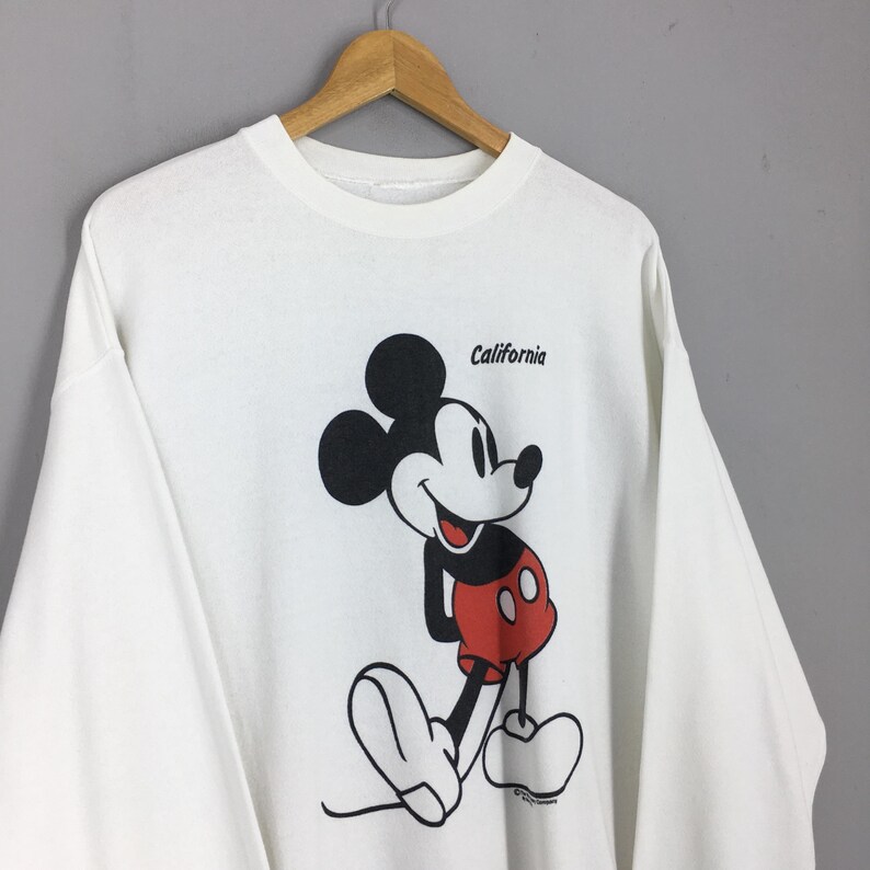 May include: White long-sleeve shirt featuring a large graphic of Mickey Mouse in black, red, and white. The word "California" is printed above the character. The shirt is hanging on a wooden hanger.