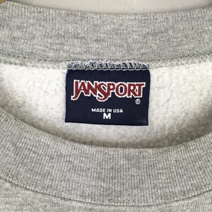 May include: Close-up of a gray sweatshirt with a navy blue tag. The tag features the brand name "JANSPORT" in white lettering, with "MADE IN USA" and the size "M" printed below.