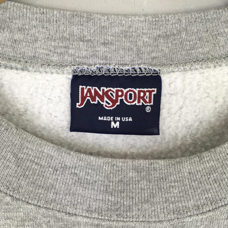May include: Close-up of a gray sweatshirt with a navy blue tag. The tag features the brand name "JANSPORT" in white lettering, with "MADE IN USA" and the size "M" printed below.