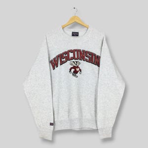 May include: A light gray sweatshirt with the word "WISCONSIN" arched across the chest in red lettering with a black outline. Below the word is a cartoon badger mascot in red and white. The sweatshirt has long sleeves and a crew neck.